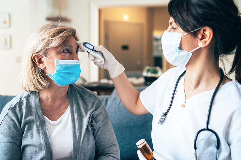 A nurse takes the temperature of a woman while both wear face masks.